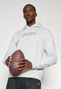 Bela kapuca z logotipom "CHIEFS" in kengurujskim žepom. Čr shorts z belimi pikami. Črn nogomet z logotipom NFL, ki ga držijo v rokah.