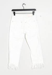 Jeans de mezclilla blanca con dobladillo deshilachado, diseño de cinco bolsillos, remaches de metal y etiqueta de parche en la cintura. Colgando de un gancho negro.
