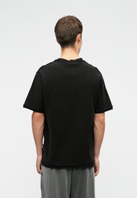 Férfi hátulról nézve, unalmas fekete, tágas, rövid ujjú pólót és laza szürke nadrágot visel egy fehér háttér előtt.