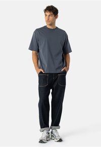 Szary krótki rękaw T-shirt o luźnym kroju, zestawiony z ciemnoniebieskimi dżinsami z widocznymi szwami i dwiema tylnymi kieszeniami, dopełniony sportowymi butami.