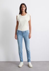 Biely rebrovaný pletený top s krátkymi volánovými rukávmi, skombinovaný so svetlomodrými slim-fit džínsy a bielymi teniskami, na neutrálnom pozadí.