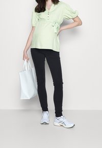 Blouse verte claire avec des manches bouffantes, boutons au niveau du col, taille nouée. Associée à un jean skinny noir et des baskets blanches. Tenant un sac fourre-tout blanc.