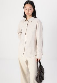 Gai bēšs, garām piedurknēm krekls ar podziņu aizdari, norādītu apkakli un teksturētu audumu. Apvienots ar gaišām biksēm un brūnu somu.