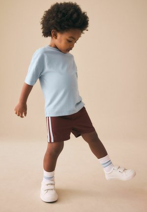 Junges Kind mit lockigem Haar trägt blaues Hemd, bordeauxrote Shorts mit Streifen und weiße Sneakers, hebt ein Bein und schaut nach unten.