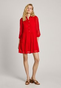 Robe rouge à boutons avec des manches transparentes trois-quarts et une jupe à volants. Matière douce et légère avec un design fluide. Sandales brunes à lanières.