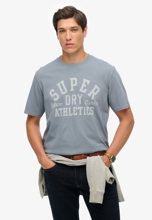 Camiseta de algodón azul claro con un estampado gráfico blanco que dice "SUPER DRY Osaka ATHLETICS". El modelo lleva jeans oscuros y un sudadera gris atada a la cintura.