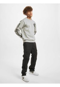 Grå sweatshirt med sort grafisk tekst på ærmerne, parret med sorte cargo-bukser. Modellen bærer hvide sneakers med guld accenter.