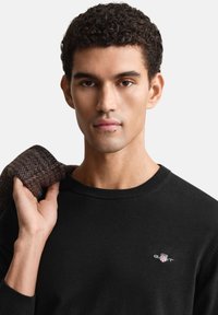 Pull noir à col rond avec un petit logo brodé sur la poitrine. Le modèle porte une veste marron texturée sur une épaule.