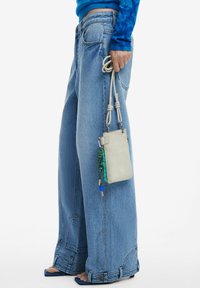 Persoon draagt wijde blauwe jeans en een blauw shirt met lange mouwen, houdt een kleine beige polstasje vast met een decoratieve touwhengsel en bedeltjes.