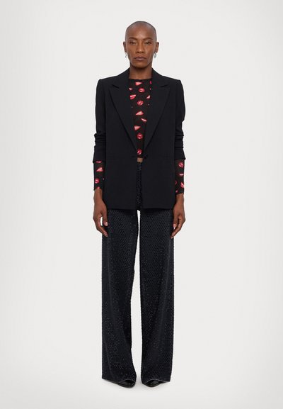 Blazer negro sobre una blusa negra estampada con un diseño de labios rojos, combinado con pantalones de pierna ancha negros con acentos brillantes.