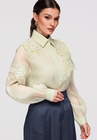 Femme portant un chemisier transparent vert pâle avec de grandes applications florales sur les épaules et les manches, associé à un pantalon taille haute bleu marine.