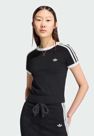 Młoda kobieta z zaplecionymi włosami nosi czarny krótki top Adidas i pasującą spódnicę z białymi paskami i logo, stojąc na jednolitym tle.