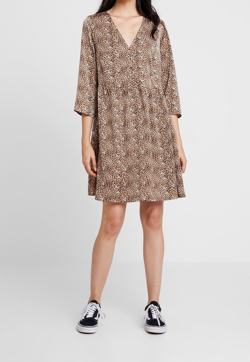 Femme portant une robe marron et noire à pois longueur genoux avec des manches trois-quarts et des baskets noires et blanches.
