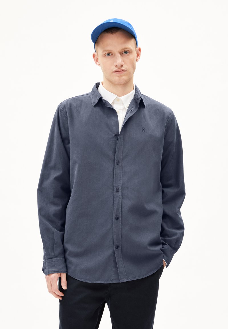 Donkerblauwe longsleeve shirt met knoopsluiting, voorzien van een witte kraag eronder en een klein logo op de borst. Gecombineerd met een blauwe pet.