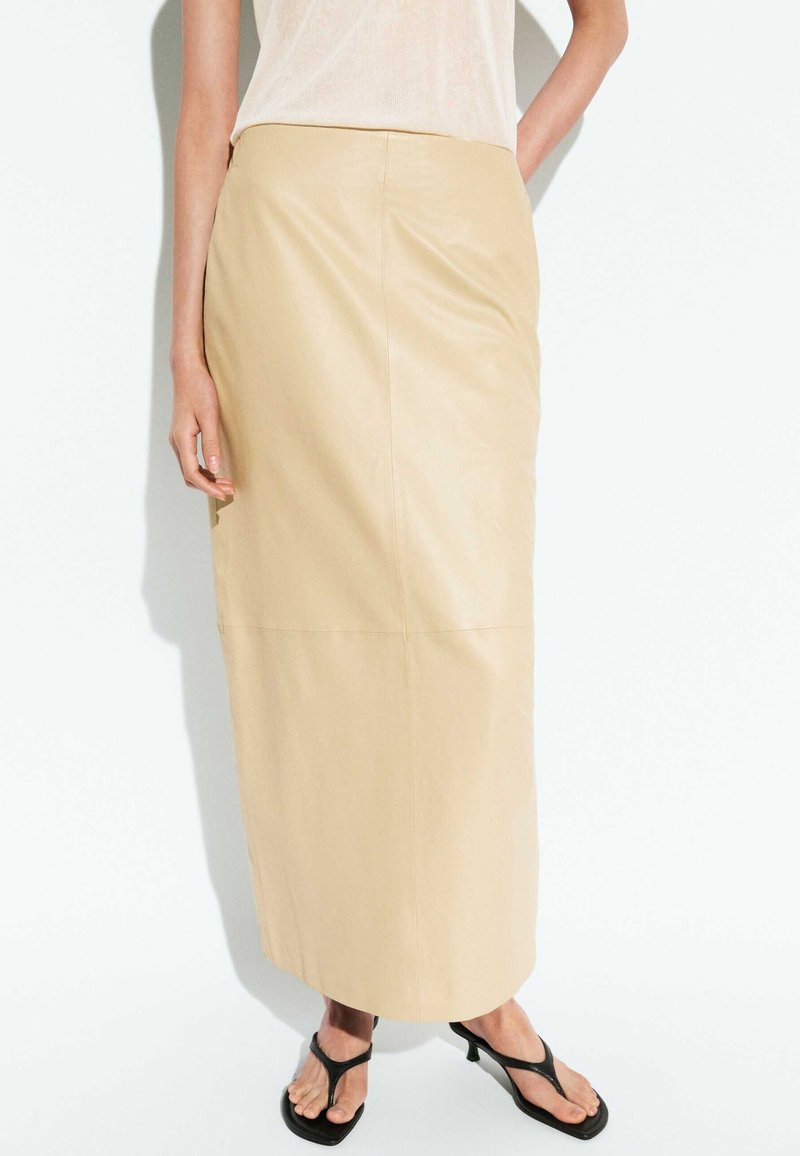 Personne portant une longue jupe beige, un haut blanc sans manches et des sandales noires à brides, debout devant un fond blanc uni.