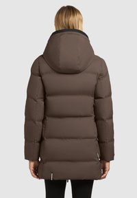Frau, die von hinten steht und einen braunen gepolsterten Wintermantel mit Kapuze sowie schwarze Leggings trägt, vor einem schlichten Hintergrund.