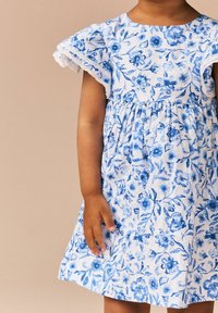 Baumwollkleid mit weißem Grund und blauen Blumenmustern, kurzen Rüschensäumen und einer gerafften Taille für einen lockeren Sitz.