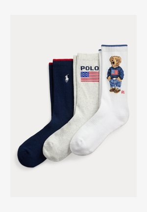 Trois chaussettes pliées en bleu marine, blanc et crème ; le bleu marine porte un logo joueur de polo blanc, la crème affiche "POLO" et le drapeau américain, la blanche a un ours avec un pull au drapeau américain.