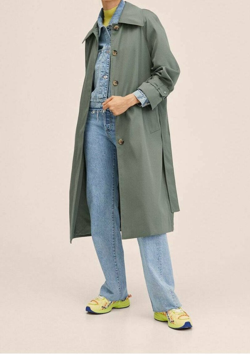 Personne portant un long manteau vert par-dessus une veste en jean bleu clair et un jean, associée à des baskets jaunes et bleues, debout devant un fond uni.