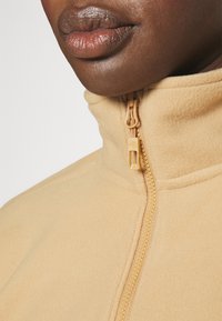 Gros plan d'une personne à la peau foncée portant une veste polaire beige zippée jusqu'au cou, montrant le curseur de la fermeture éclair et la texture du tissu.