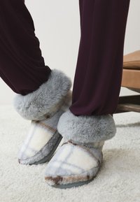 Graue und cremefarbene Plüschpantoffeln mit einer Kunstpelzbündchen, mit einem geometrischen Muster und einer strukturierten Oberfläche, kombiniert mit dunkelvioletten Hosen.
