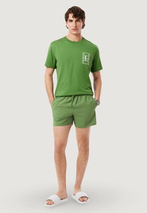 Ung mand stående med hænderne i lommerne iført en grøn t-shirt, grønne shorts, hvide slidesandaletter og et armbåndsur mod en lys baggrund.