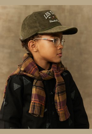 Oliwkowy kapelusz z korduroju z haftowanym napisem, okulary i woskowany wełniany szalik w kolorach żółtym, burgundowym i czarnym, nałożony na czarną zapinaną kurtkę.