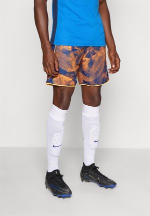 Blå sports t-shirt, orange og marineblå mønstrede shorts med gul kant, hvide sokker, sorte fodboldstøvler med blå accenter.