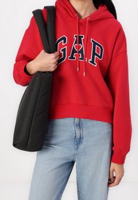Czerwona bluza z kapturem z logo GAP, ściągaczem przy kapturze i ściągaczem u dołu. W zestawie z jasnoniebieskimi dżinsami oraz czarną pikowaną torbą na ramię.