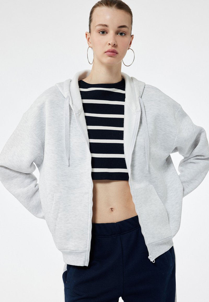 Lys grå zip-up hoodie med en frontlomme, har en stor hætte og bæres over en marineblå stribet cropped top. Enkel, afslappet design.