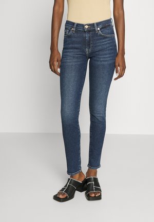 Kvinde iført mørkeblå skinny jeans, beige ærmeløs top og sorte åbne blokhælede sandaler med hvide stikninger, stående mod en hvid baggrund.
