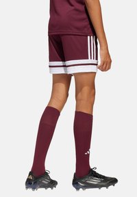 Bordeauxrotes Fußballtrikot mit weißem Einsatz; die Shorts haben zwei Streifen, hohe Socken in Bordeauxrot. Schwarze Fußballschuhe mit hellen Akzenten und strukturiertem Profil.