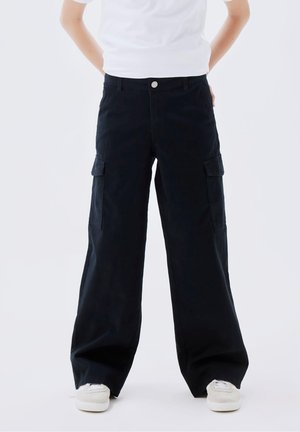 Pantalon cargo à jambes larges en coton noir, avec poches latérales, fermeture à bouton, et une coupe décontractée, associé à des baskets blanches.