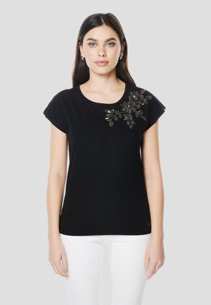 Camiseta negra de manga corta con escote redondo, con detallados adornos de cuentas florales en el hombro izquierdo delantero.