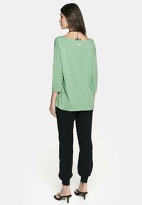 Luźny, zielony top z bawełny o szerokim dekolcie i rękawach do trzech kwart, zestawiony z czarnymi spodniami zwężającymi się ku dołowi i sandałami na obcasie.