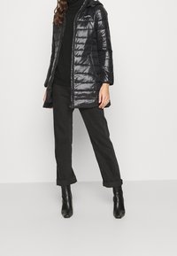 Chaqueta acolchada negra y brillante con capucha y cierre de cremallera. Cuenta con costuras horizontales y un logo en el pecho. Combinada con pantalones y botas negros.