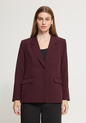 Femme aux cheveux raides châtain clair portant des boucles d’oreilles argentées en forme d’anneaux et un blazer violet foncé par-dessus un haut et un pantalon noirs, debout devant un fond uni.