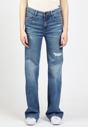 Woman wearing blue distressed straight-leg jeans and white sneakers, standing against a plain white background.