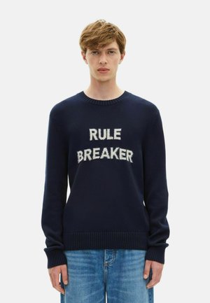 Pull marin en tissu tricoté arborant le texte "RULE BREAKER" en lettres blanches duveteuses. Col et poignets côtelés. Coupe décontractée.