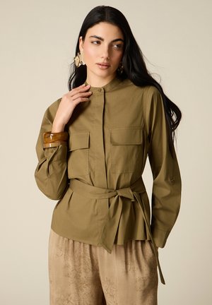 Donna con capelli lunghi e scuri che indossa orecchini grandi dorati, una camicia verde oliva con cintura e tasche frontali, e pantaloni beige con motivo floreale.