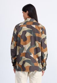 Tarn- und olivgrünes Camouflage-Hemd mit lockerer Passform. Zu den Hauptmerkmalen gehören lange Ärmel, ein Kragen und ein abgerundeter Saum.