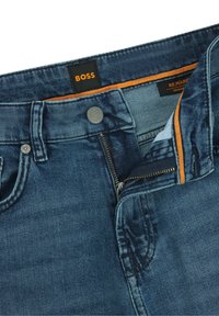 Pantalones vaqueros de mezclilla azul oscuro con cierre de cremallera estándar, cierre con botón de metal y un detalle en contraste de color naranja a lo largo de la pretina.