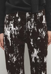 Pantalon motif acid wash noir et blanc porté avec un haut noir à manches longues, mains détendues le long du corps.
