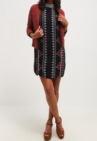 Kvinna som bär en mönstrad svart klänning med en vinröd cardigan och bruna platåskor, poserar självsäkert med händerna på höfterna.