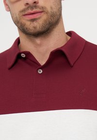 Άνδρας με γένια που φοράει μια μπλούζα πόλο σε μπλοκ χρωμάτων μπορντό και λευκό, με γιακά και δύο κουμπιά κουμπωμένα.