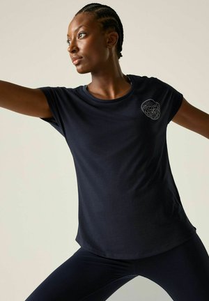 Woman with braided hair wearing a black t-shirt and leggings stretching her arms in a light-colored room.