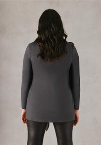 Camiseta de manga larga, gris, con una textura suave y un corte holgado, combinada con leggins negros, brillantes. La camiseta tiene un dobladillo redondeado.