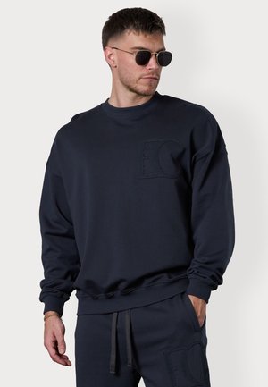 Sweatshirt bleu marine avec col rond, manches longues et un logo "EC" texturé sur la poitrine. Tissu doux et détails des poignets et de l'ourlet côtelés.