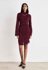 Burgundy lace dress with long sleeves, a fitted silhouette, a scalloped hem, and a textured floral pattern, paired with black pointed flats.