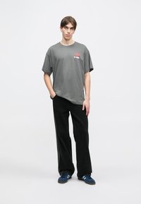 Pelēks kokvilnas T-krekls ar īsām piedurknēm, kas rotāts ar sarkanu logo, apvienots ar melniem platām biksēm un ziliem kedām ar trim svītrām.